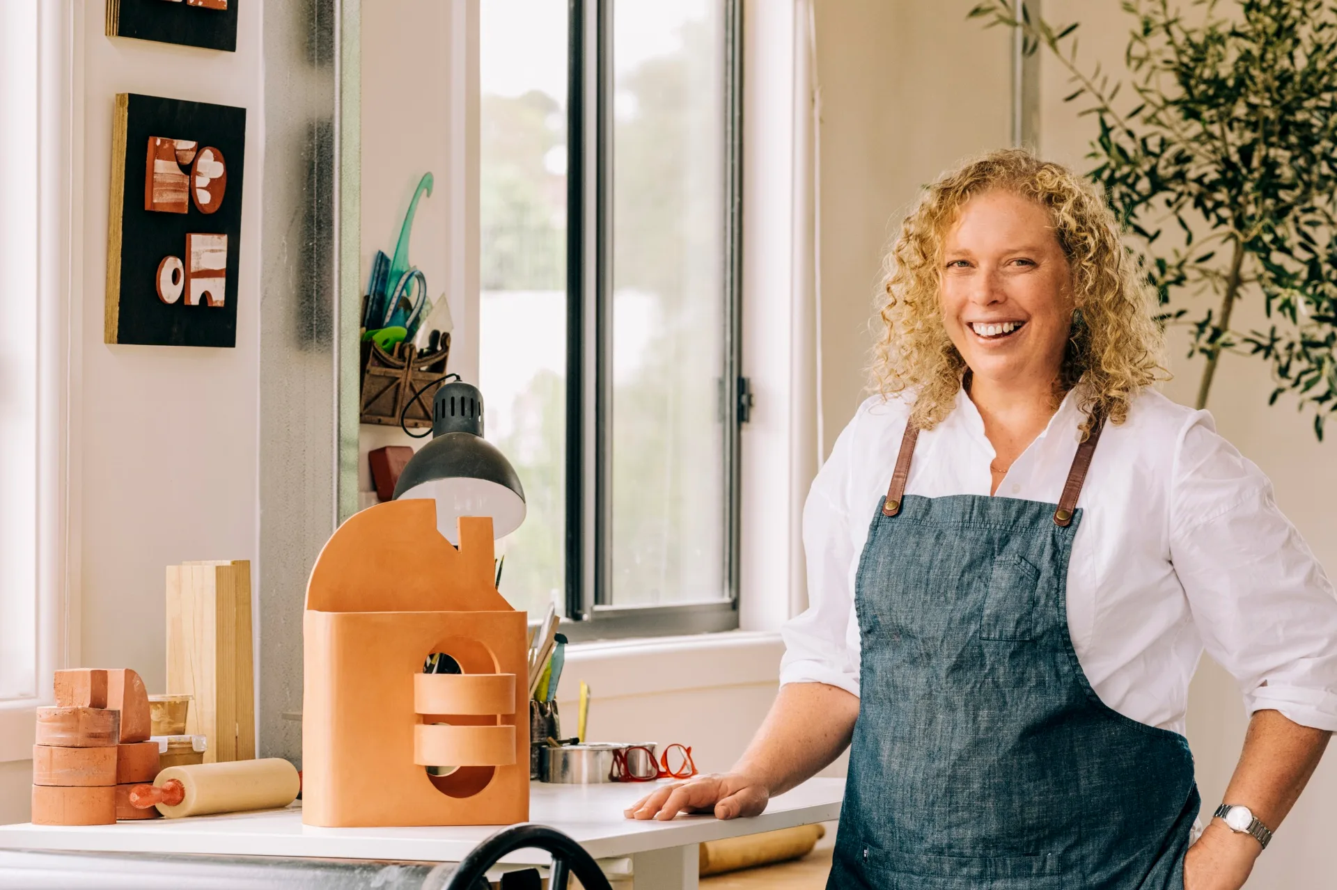 Ceramic artist in apron smiling in studio with clay creations on table.