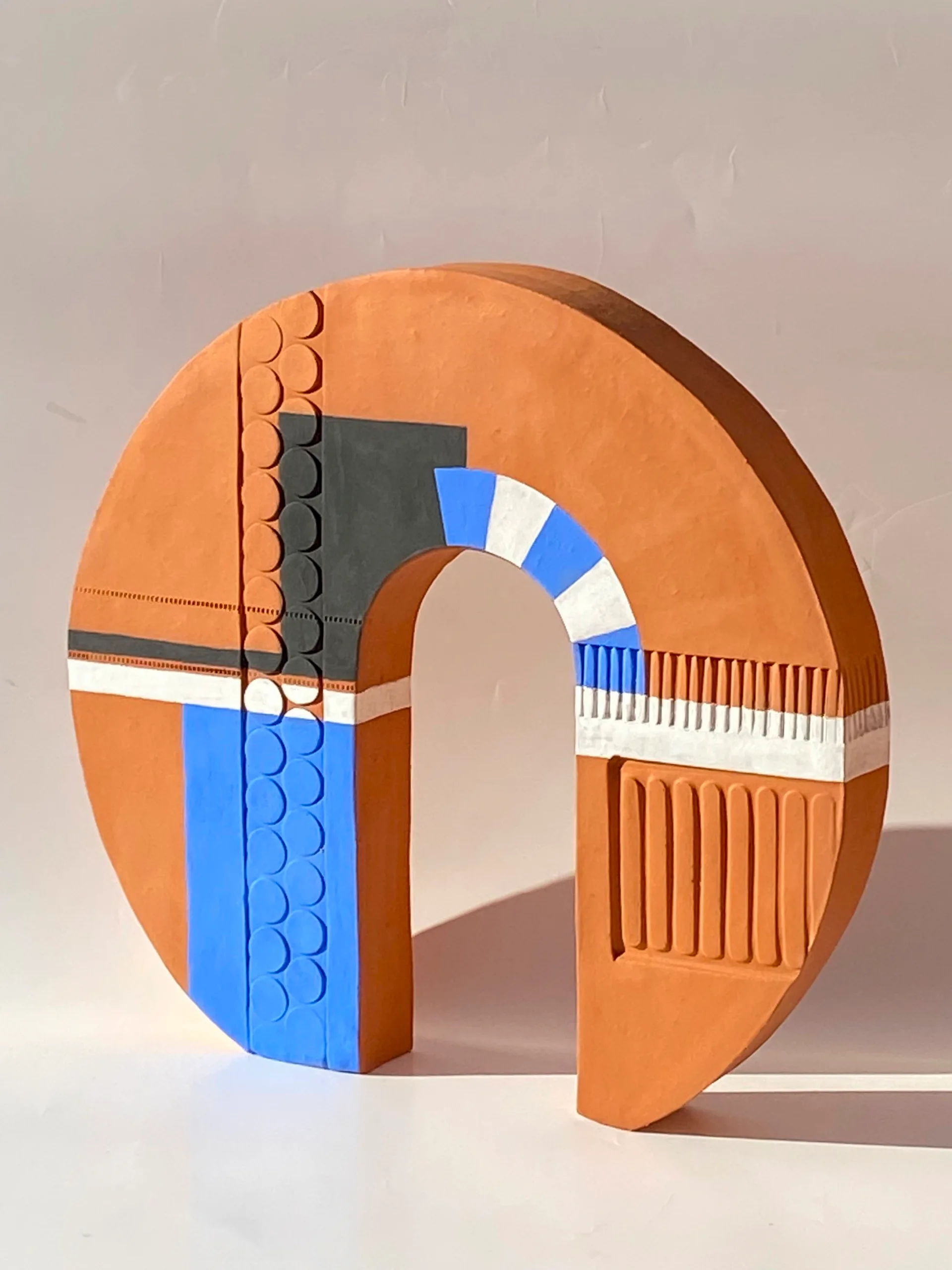 A clay abstract sculpture with a round shape and an arched void in the middle.