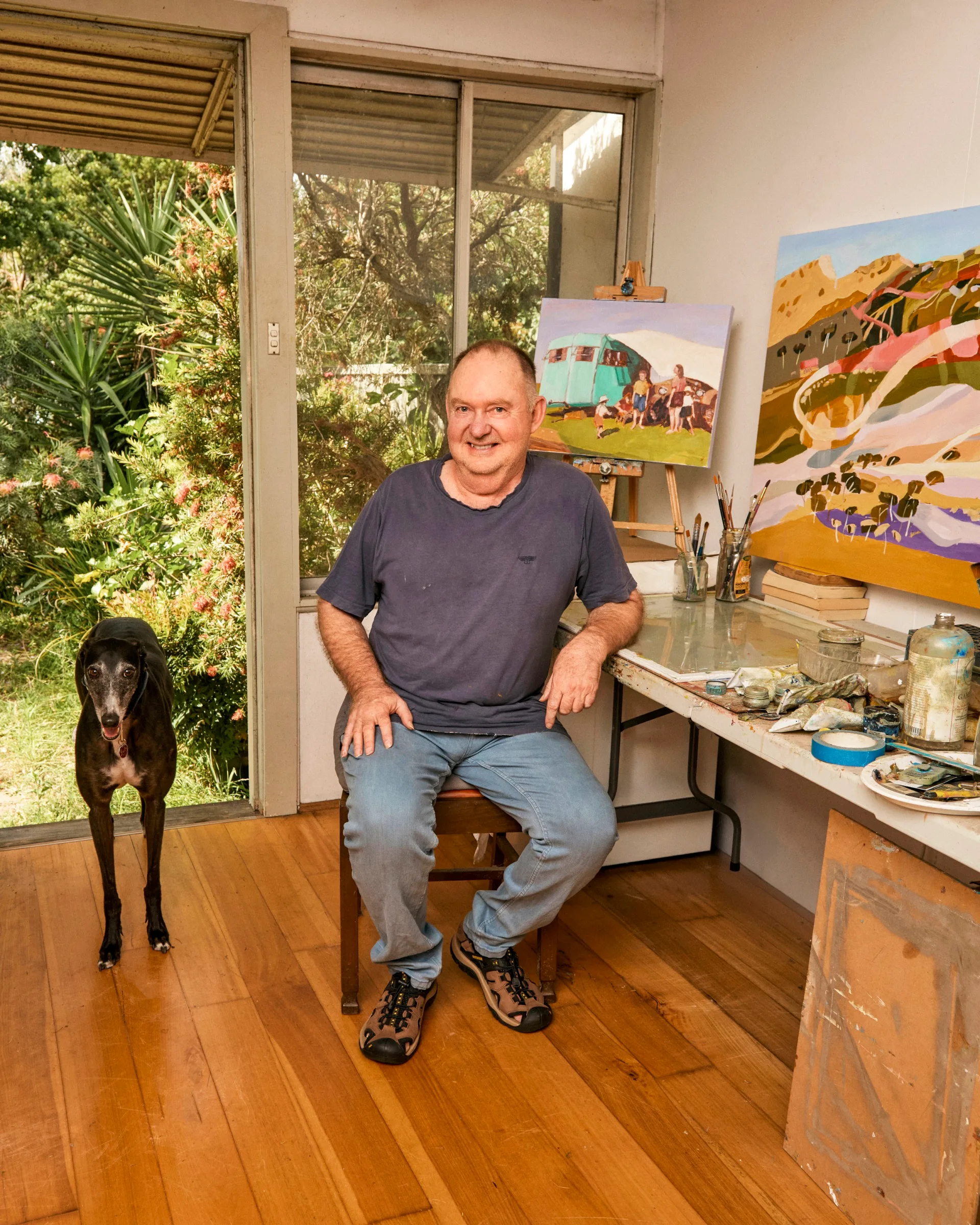 Man sitting in an art studio with paintings around, a black dog standing nearby, large window in the background.