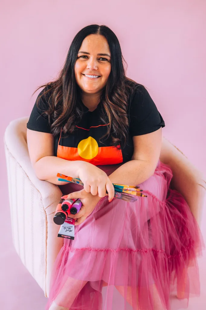 Smiling woman in a pink skirt and black t-shirt with the Aboriginal flag on it, holding paintbrushes and paint tubes, sitting against a pink background.