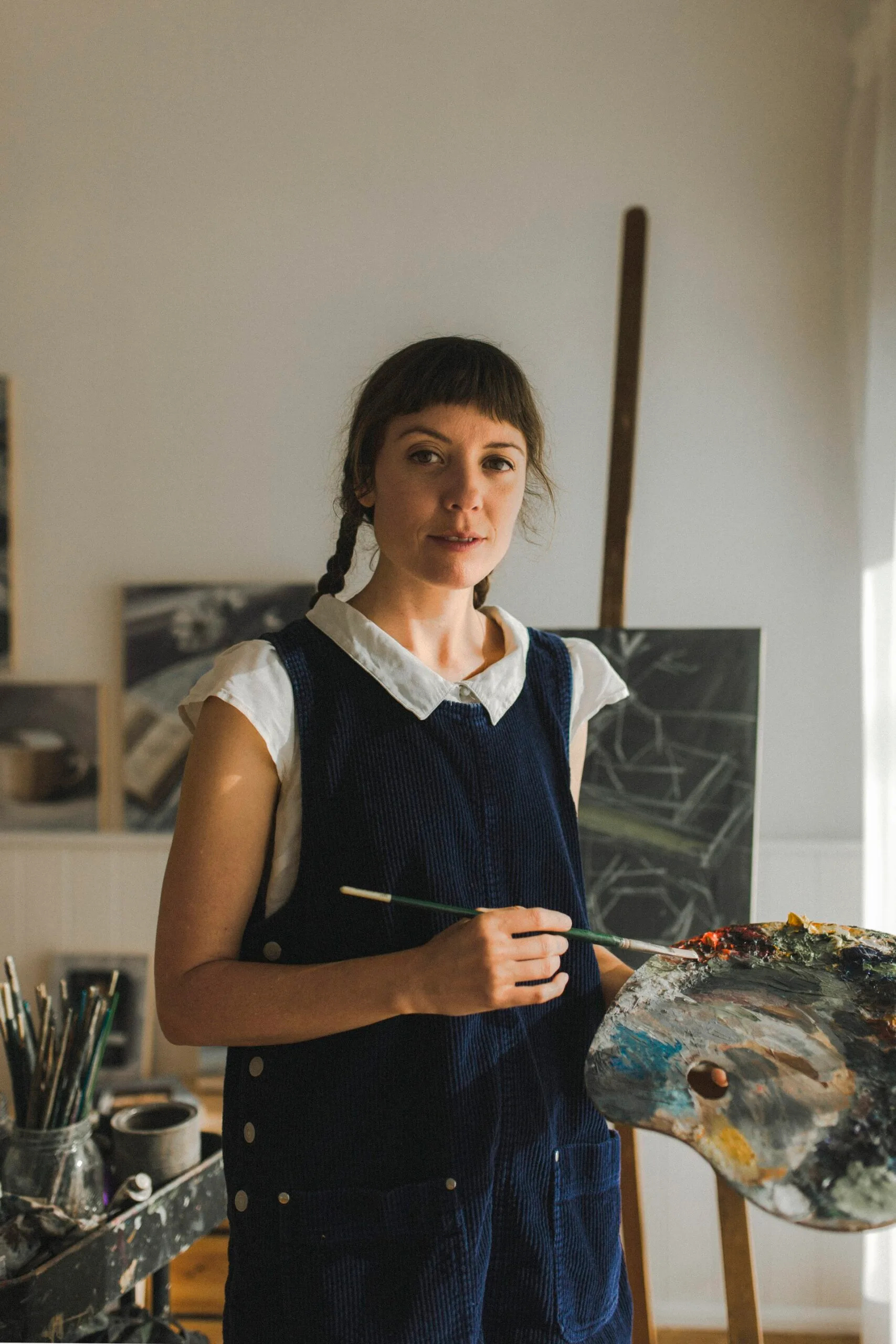 Woman with pigtails holding a paintbrush and palette in an art studio, standing near brushes and paintings.