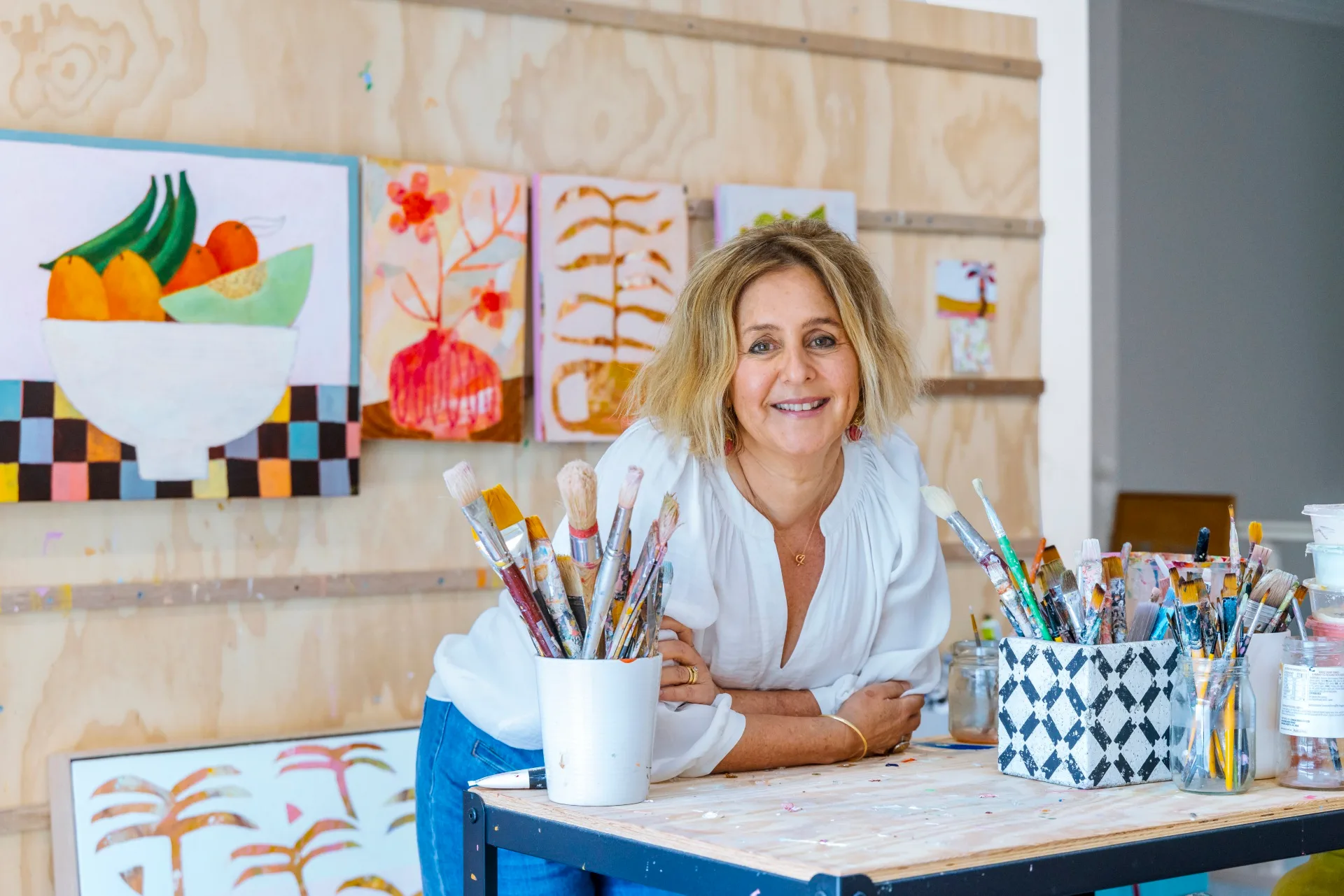 Artist in a bright studio, smiling, surrounded by colorful artwork and paintbrushes.
