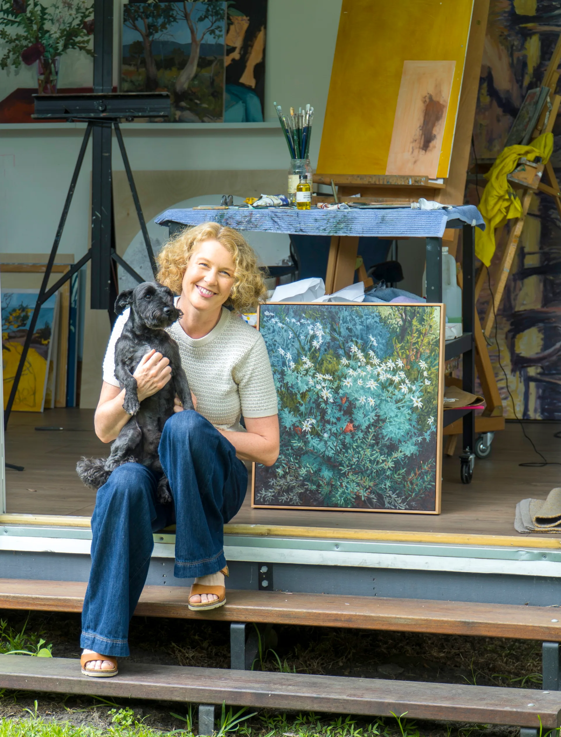 Artist sits on steps holding a dog, with a painting studio behind her.