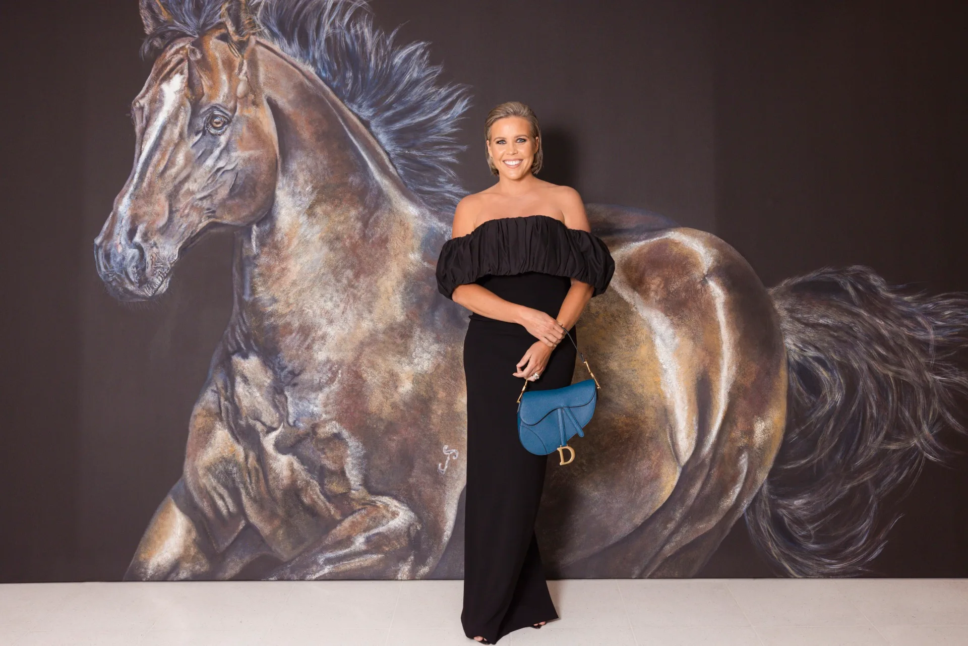 A woman in a black dress stands in front of a large horse painting, holding a blue handbag