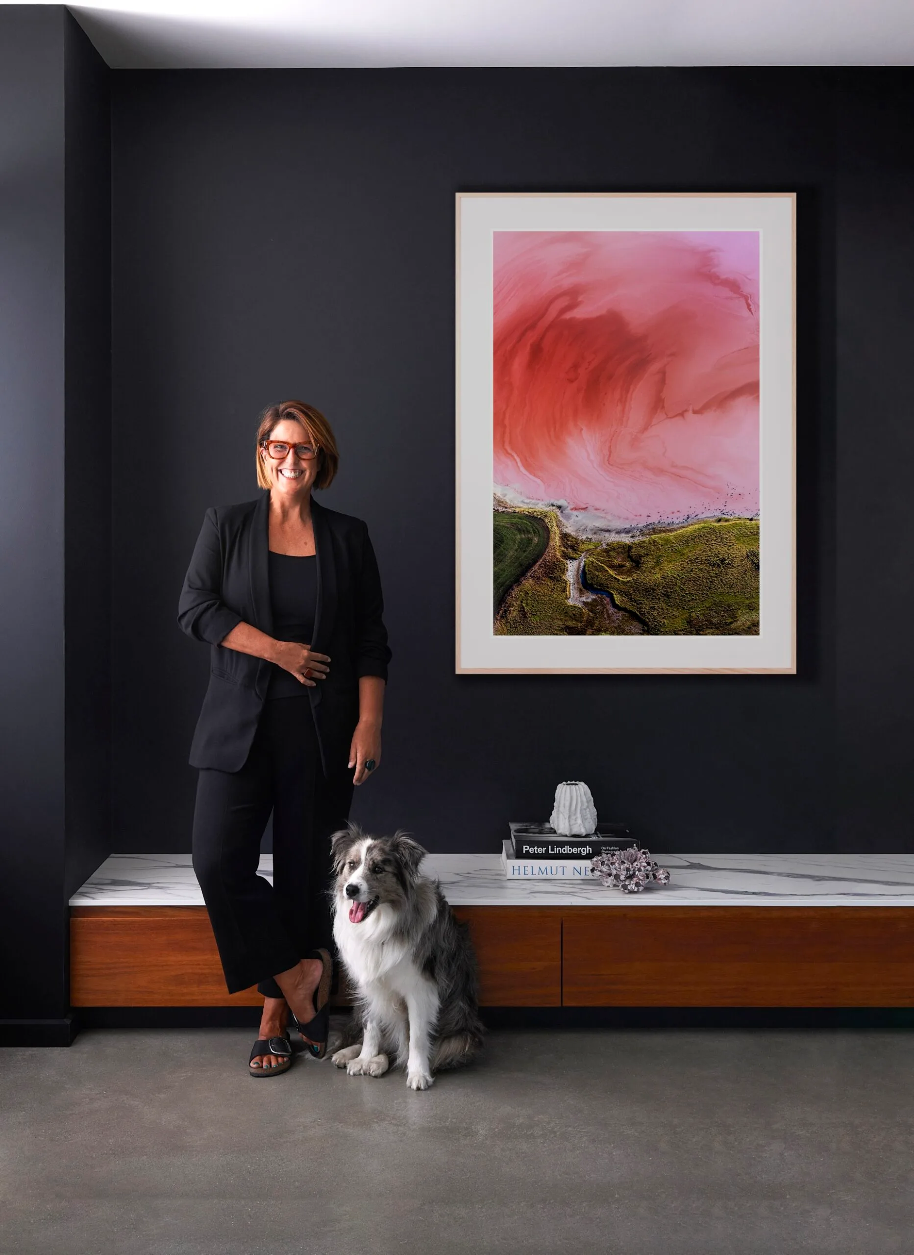 A person in black with glasses stands next to a seated dog, in front of a large pink art piece on a dark wall.