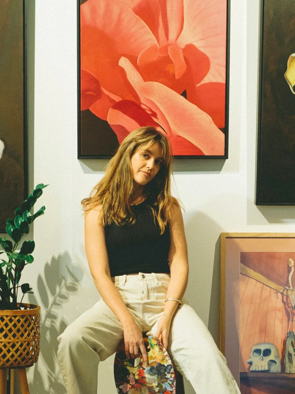Artist sitting with a paint palette in front of vibrant floral paintings in a gallery setting.