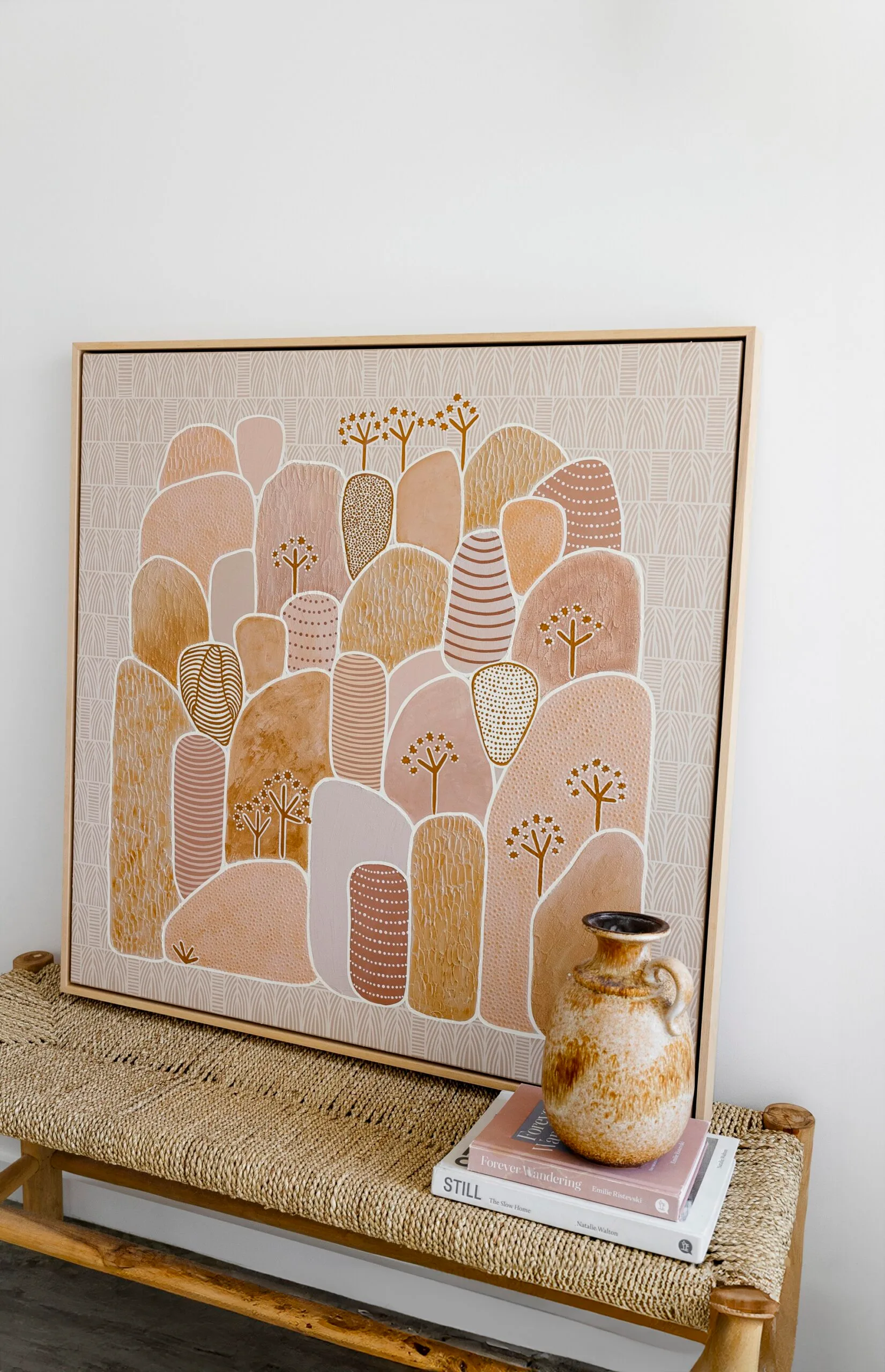 Framed Indigenous artwork with earth tones on a woven bench, accompanied by a ceramic vase and stacked books.