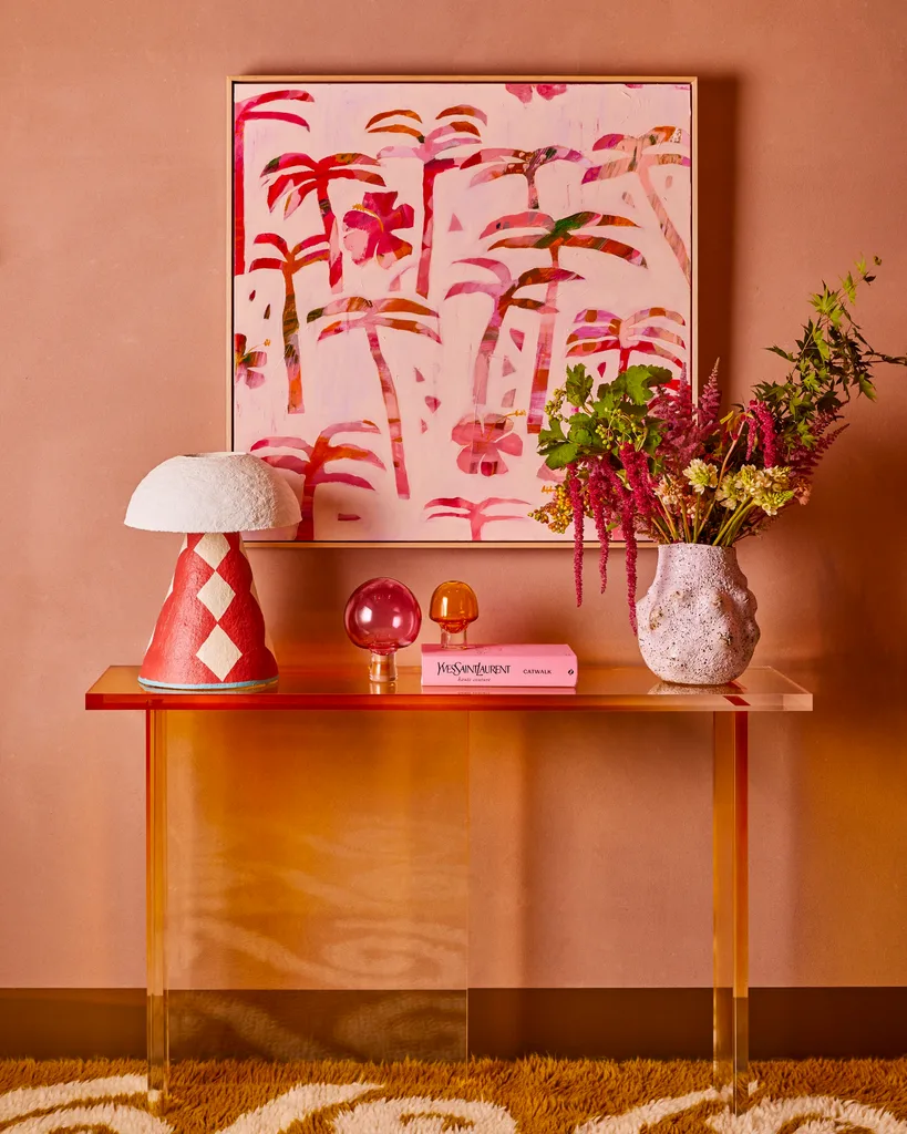 Colorful console table with art featuring palm tree silhouettes in pink shades, decorative lamps, a pink book, and a vase of flowers against a warm-toned wall.