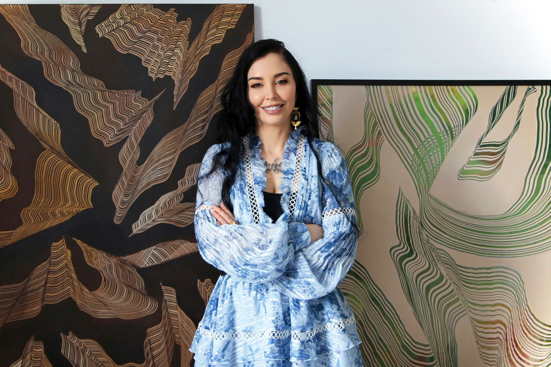 Woman in blue dress stands smiling with arms crossed, framed by abstract artwork on a wall.