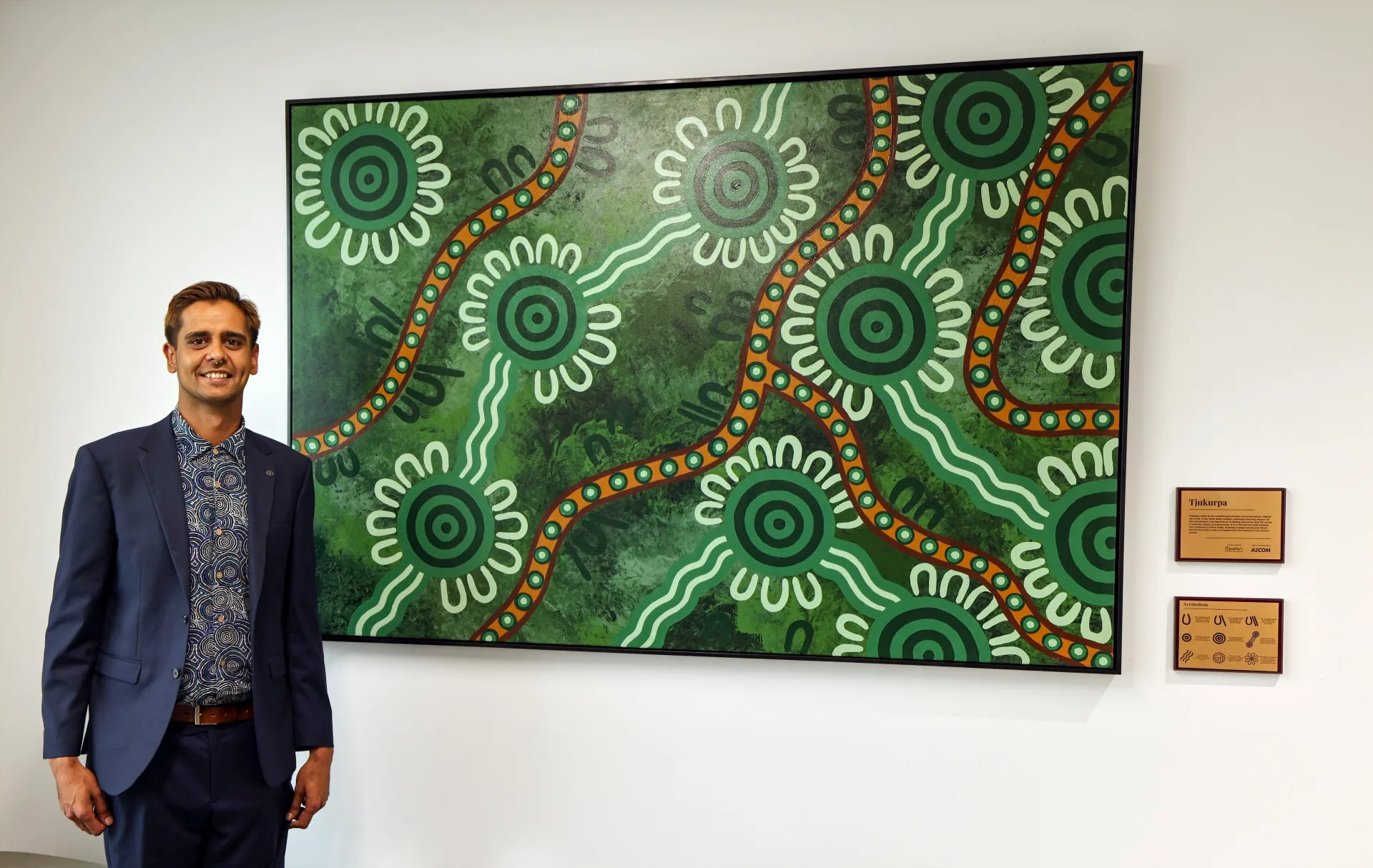 An artist stands next to a large Indigenous painting featuring lots of green colours and lines of orange paint.