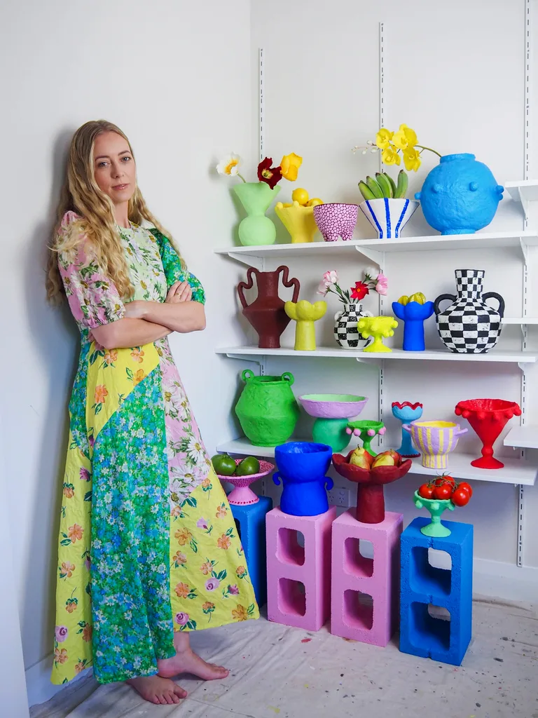 A woman in a colorful dress stands by shelves displaying bright, decorative vases and fruit.