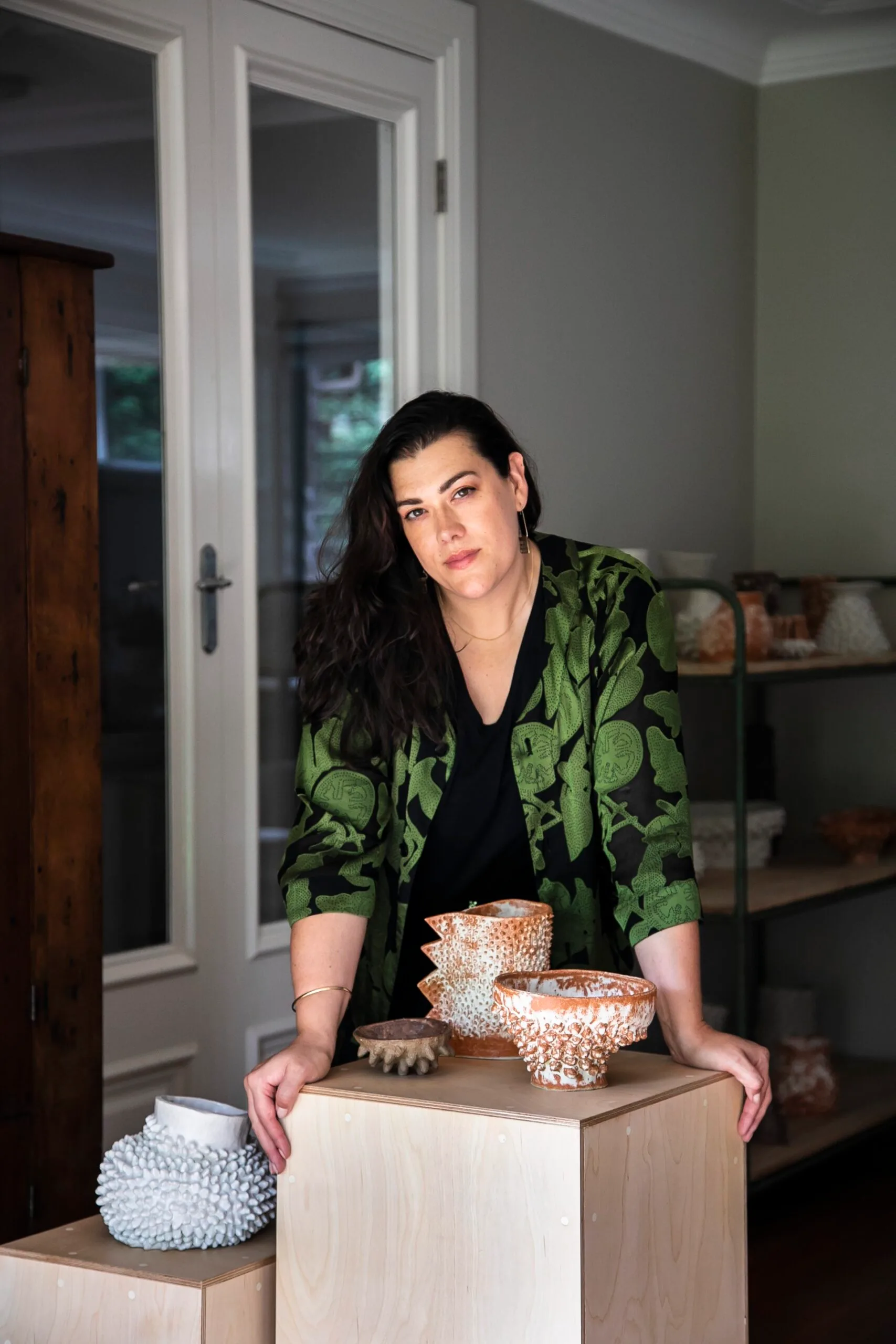A woman in a green jacket leans on a wooden table displaying textured ceramic pots