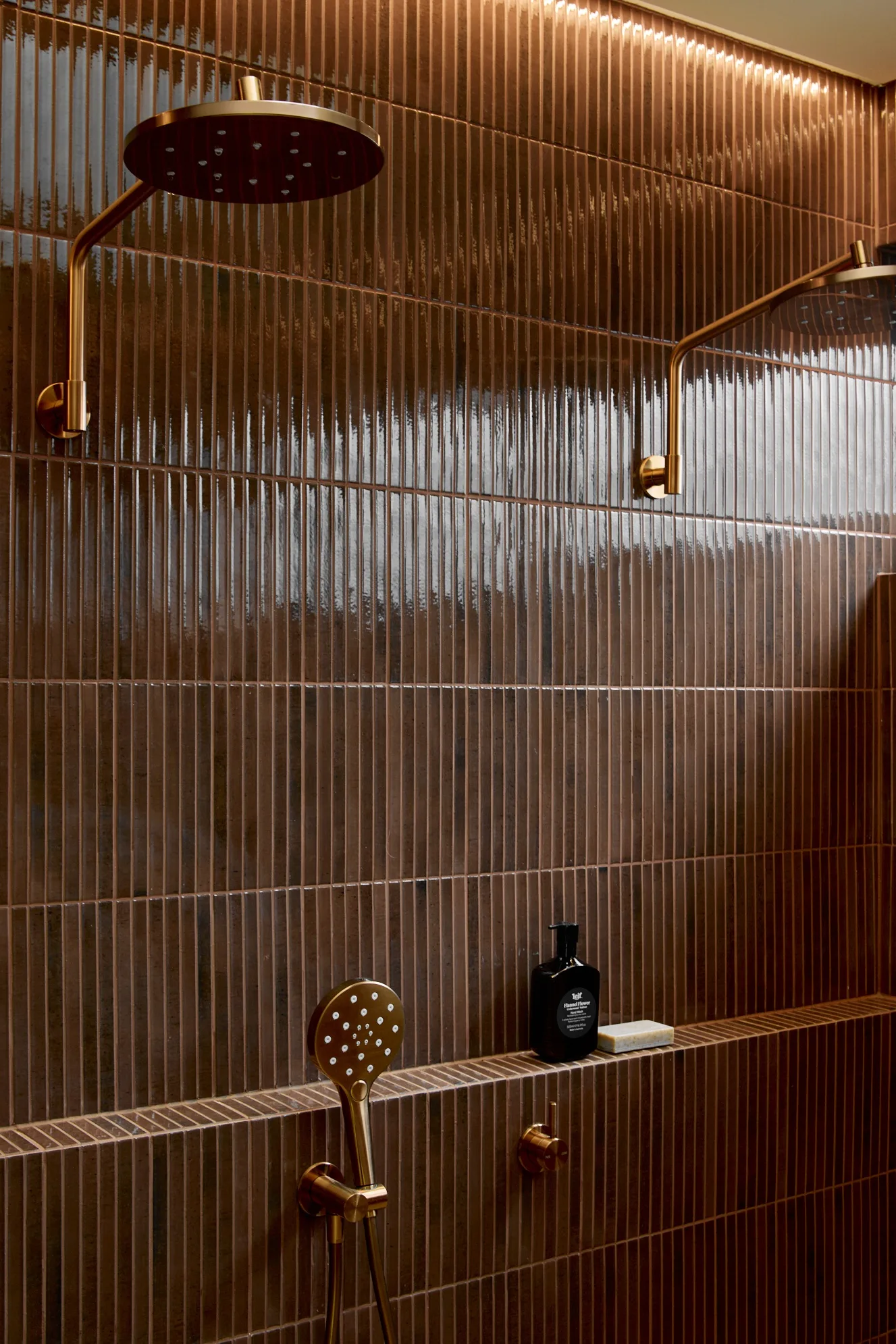 Close up of a double shower, with two shower heads and one handheld shower head, on walls finished in burgundy kit-kat wall tiles. The shower hardware is Brushed Bronze.