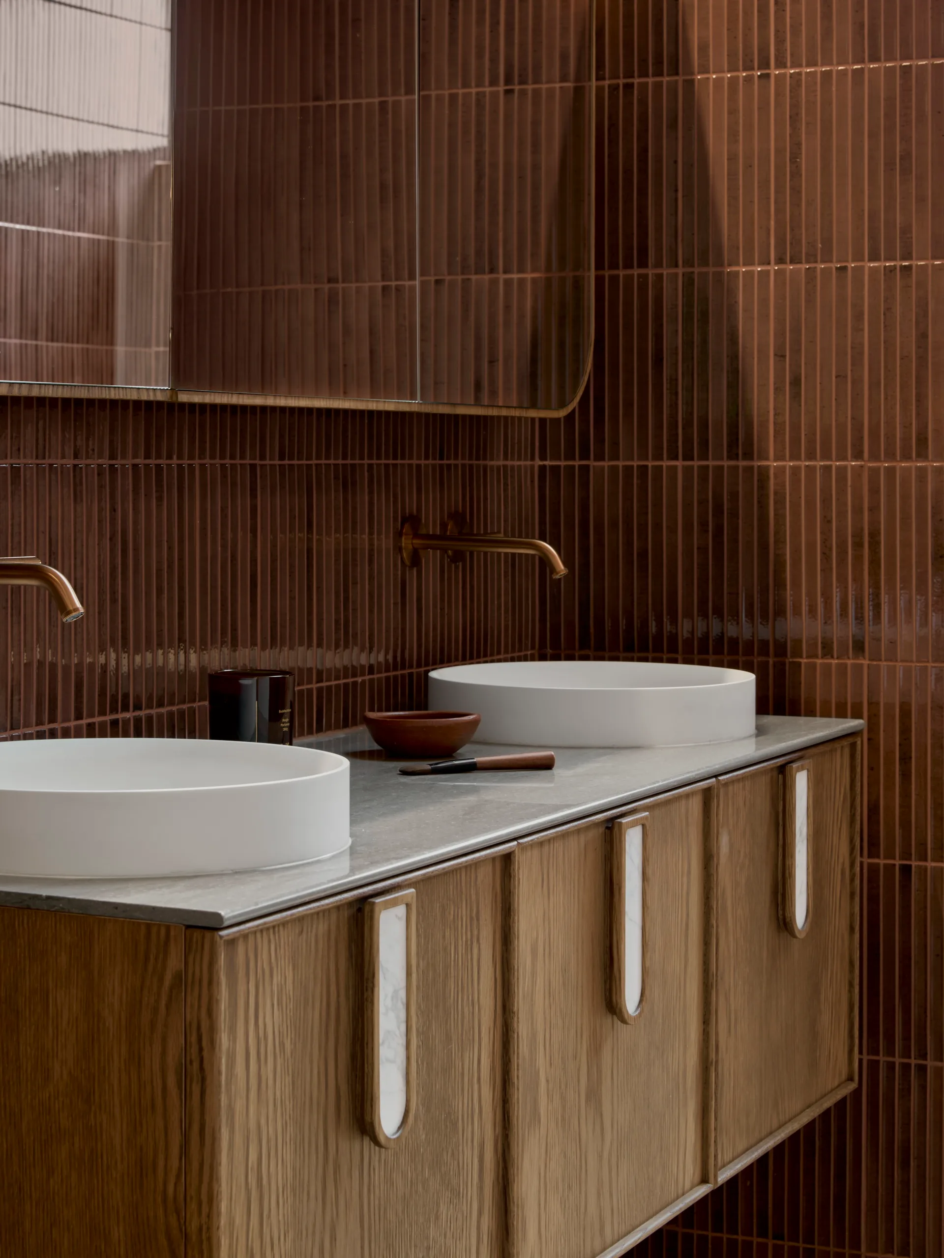 Elegant bathroom vanity with wooden vanity, two round basins on engineered stone benchtop, burgundy gloss kit-kat wall tiles, and brushed bronze faucets.