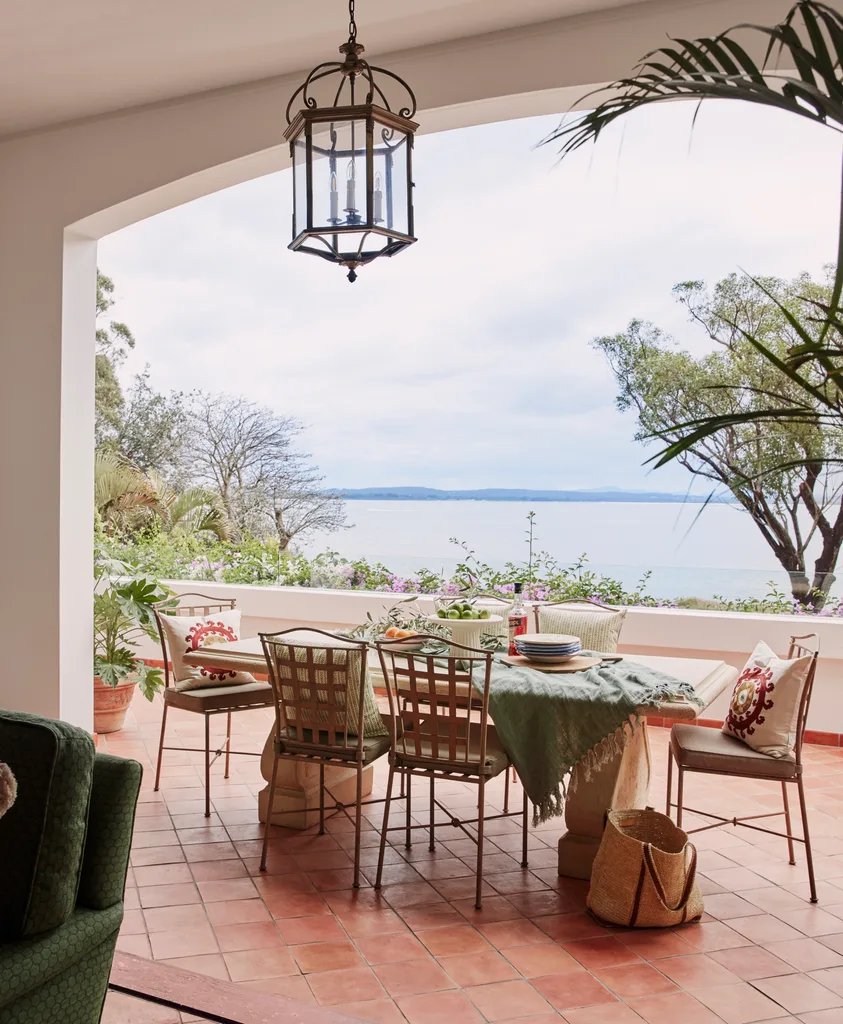 Covered patio with dining table, chairs, potted plants, overlooking a scenic coastal view. Hanging lantern above.