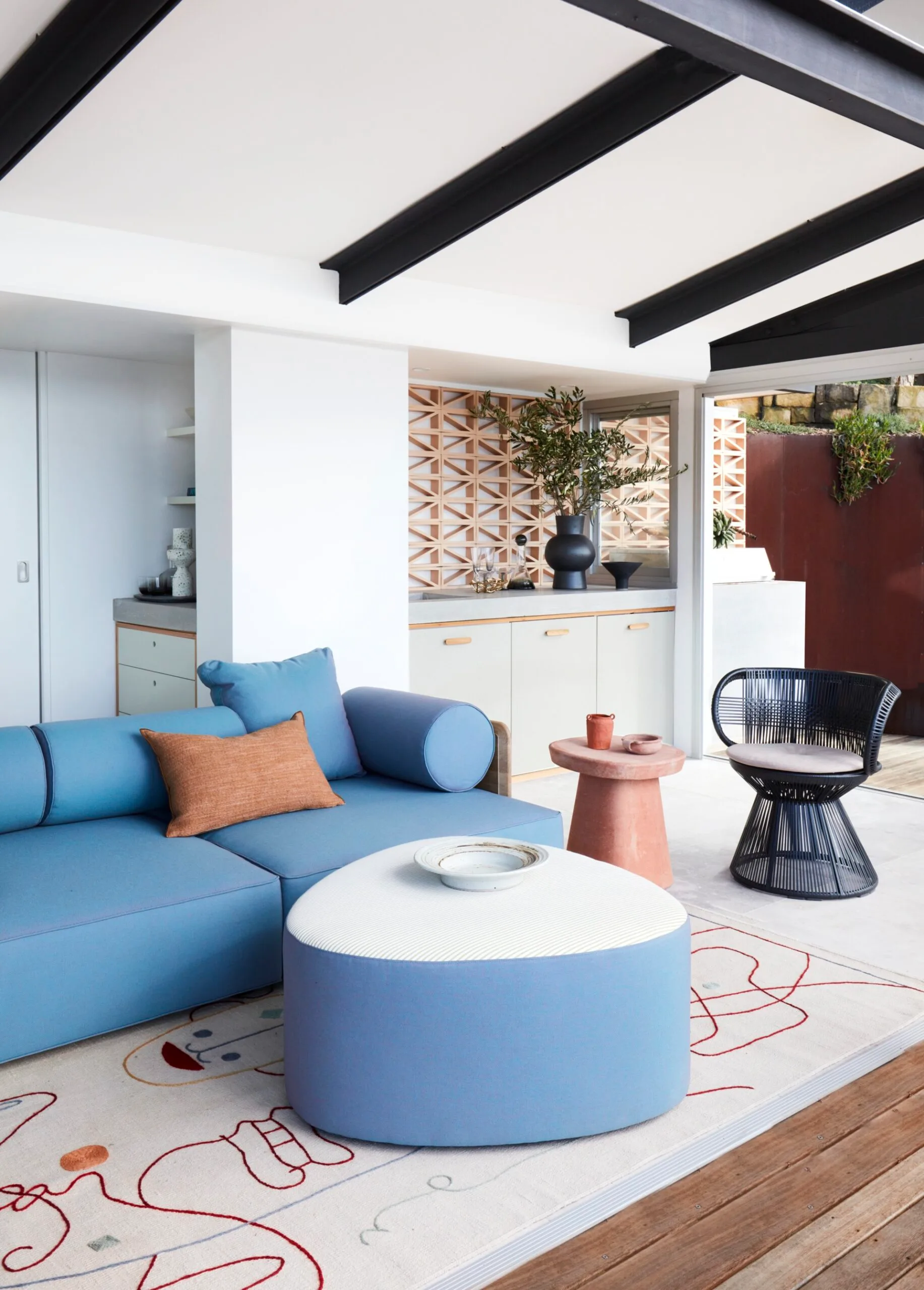Stylish alfresco living room with blue sofa, matching curved blue ottoman with a white top, patterned rug, and modern decor featuring plants and a round side table.