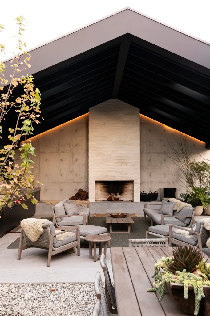 Modern outdoor living area with a vaulted roof, cozy seating, central fireplace, and surrounding plants.