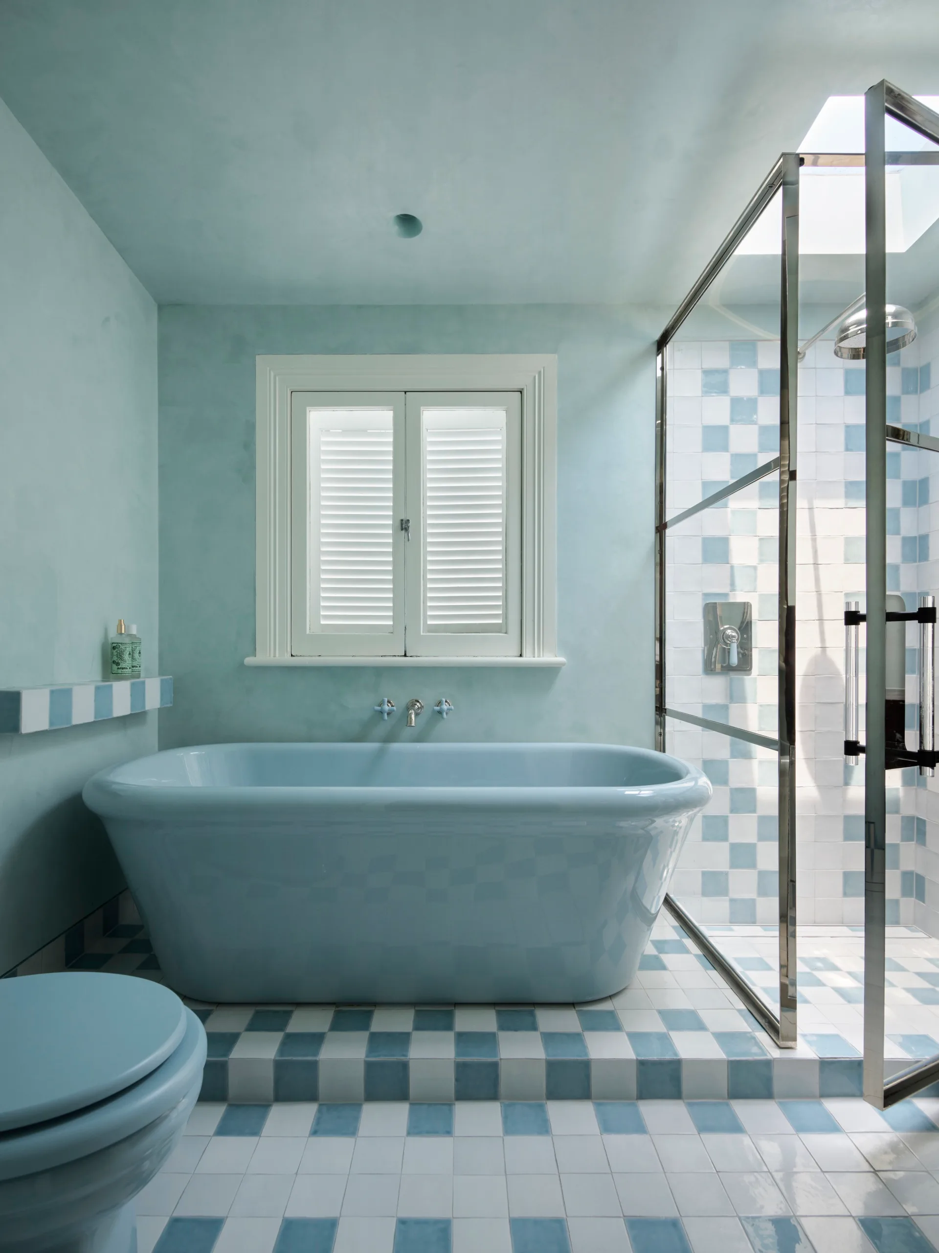 Luxurious light blue bathroom with a freestanding blue bath tub, glass shower doors, blue and white checkered tiles, and a window with shutters.