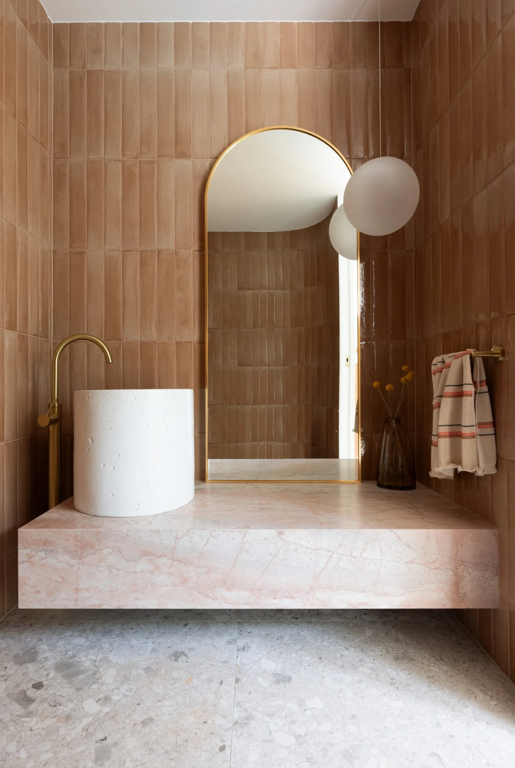 Contemporary bathroom with pink marble countertop, beige tiles, gold faucet over a pedestal basin that is round, arched mirror, and striped towel.