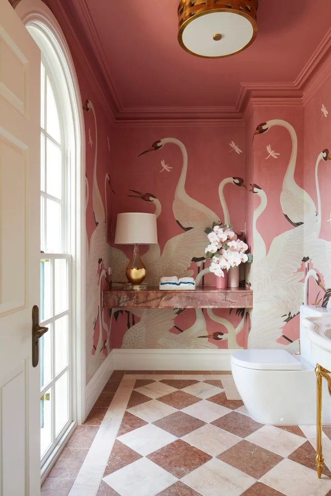 Pink bathroom with elegant crane wallpaper, checkered marble floor, wall-mounted shelf, lamp, floral decor, and white toilet.