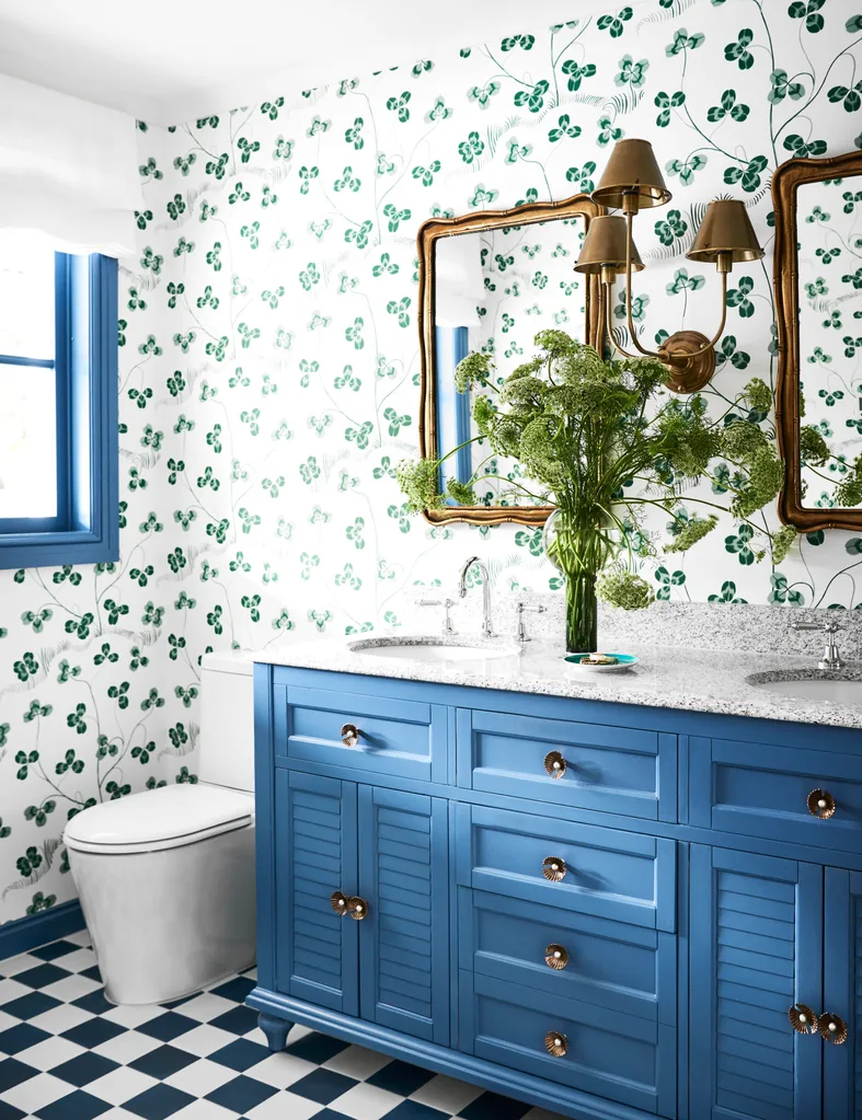 Bathroom with blue vanity, floral wallpaper, brass fixtures, and a vase filled with greenery on countertop. The floor is checkered blue and white.