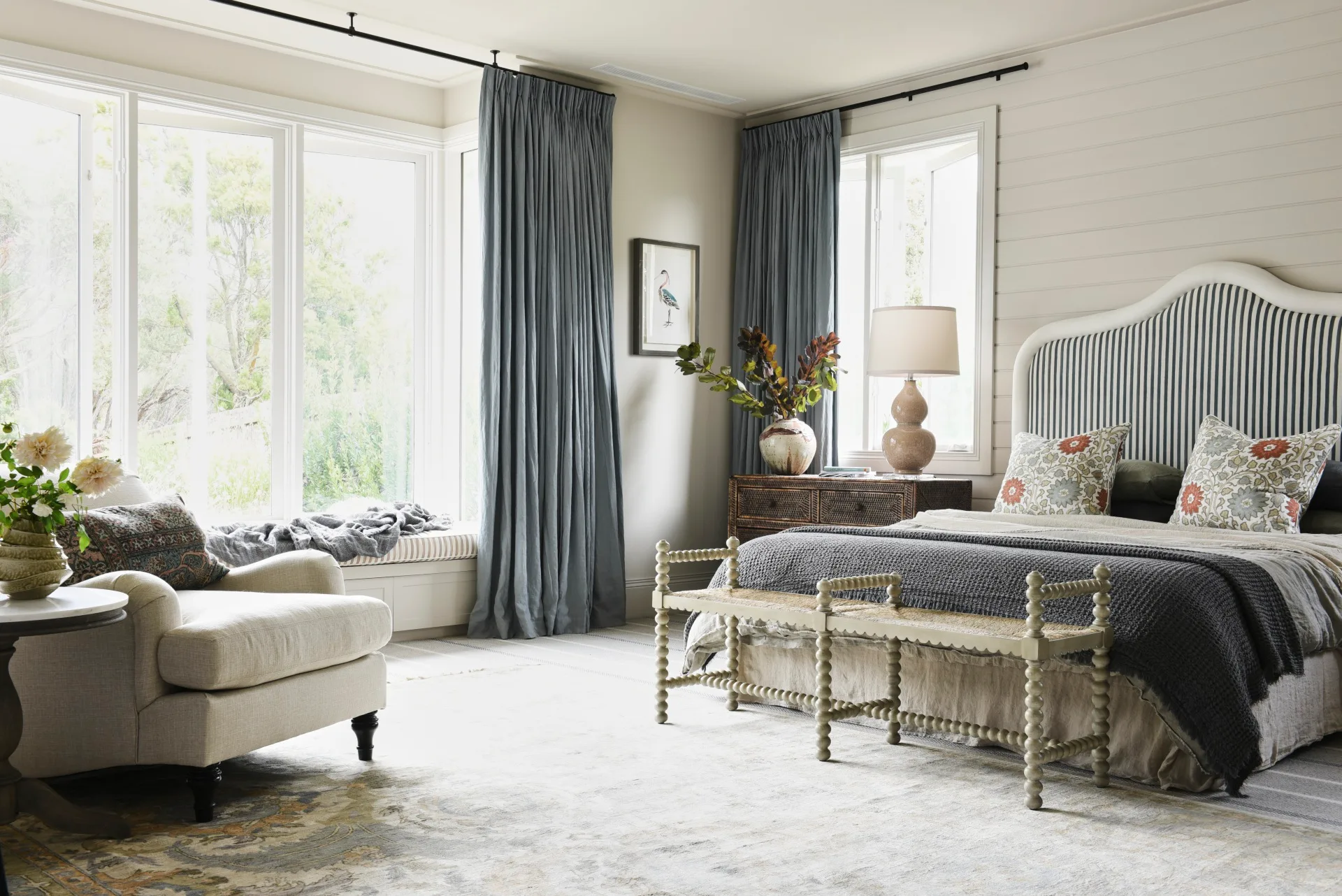 Elegant bedroom with a large bed, striped headboard, floral pillows, armchair, and large windows with blue curtains.