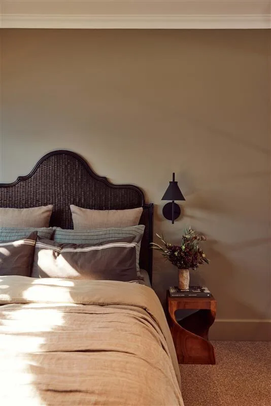 Bedroom with a dark wicker headboard, beige bedding, side table, vase with flowers, and a black wall sconce.