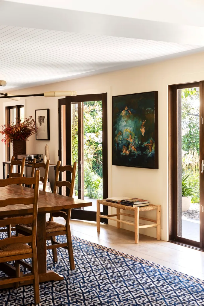 Dining room with wooden table, wooden dining chairs, large painting, books on bench, glass doors, and blue patterned rug.