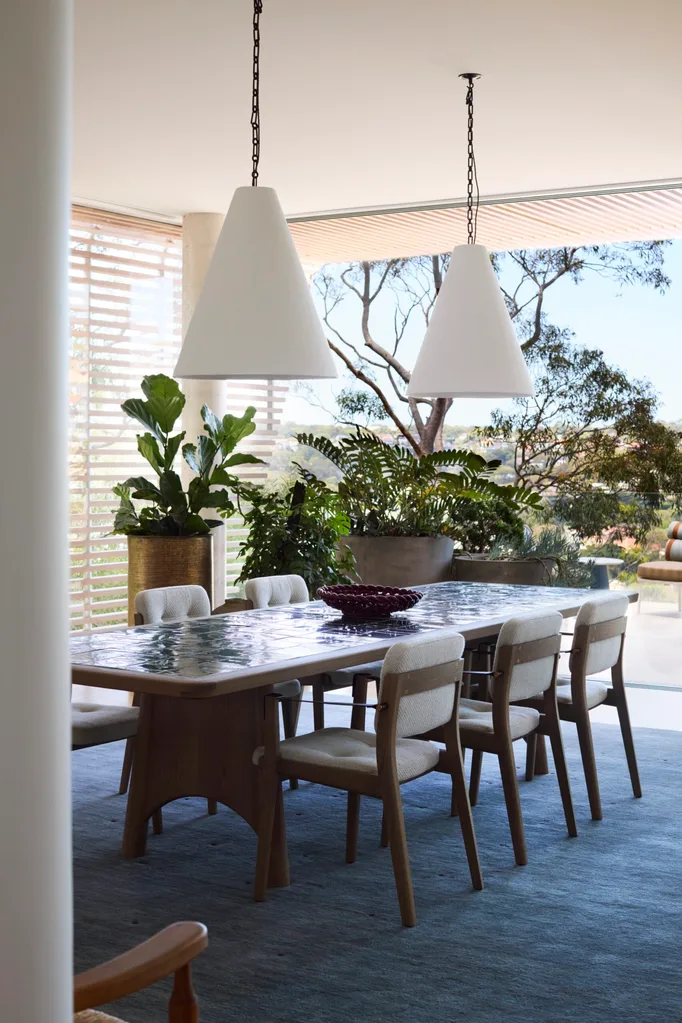 Dining room with a long table, cushioned chairs, two pendant lights, large indoor plants, and a view of trees outside. The back of the space opens up to create easy indoor-outdoor flow.