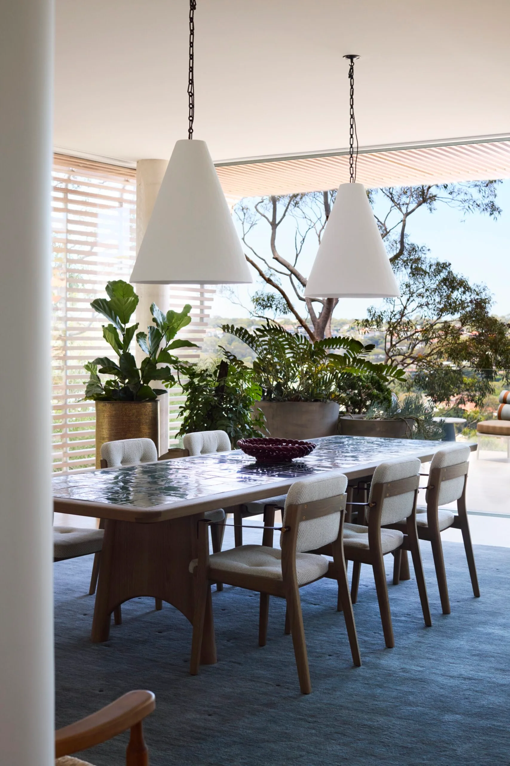 Dining room with a long table, cushioned chairs, two pendant lights, large indoor plants, and a view of trees outside. The back of the space opens up to create easy indoor-outdoor flow.