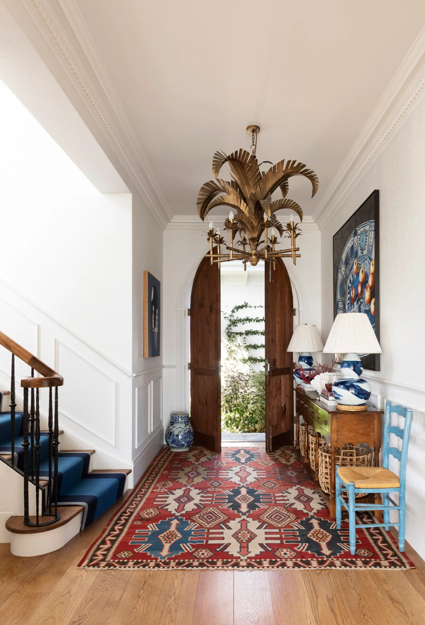 Classical entryway with elaborate chandelier in the shape of palm tree fronds, open arched wooden doors, colorful rug, and decorative side table with lamps.