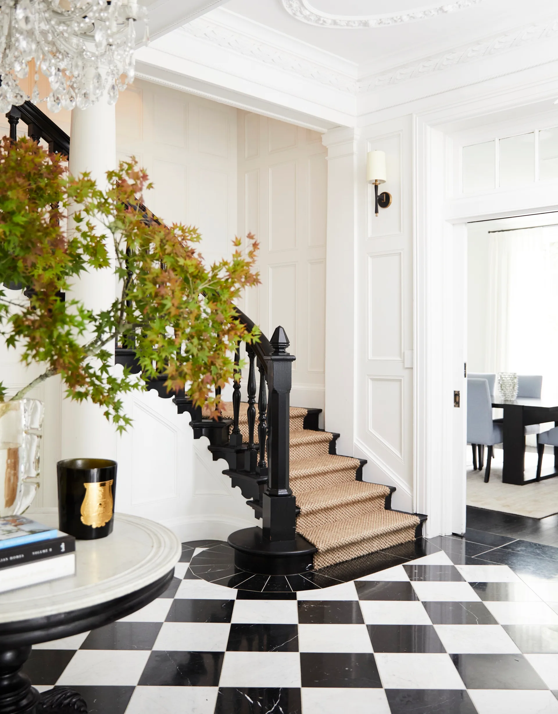 Elegant curved staircase with black railing, checkerboard floor, chandelier, and a table with a plant in a bright foyer. The walls are white with elegant and ornate moulding.