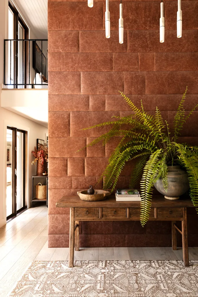 Brown textured wall, wooden table with books and a potted fern, pendant lights above, large patterned rug below.