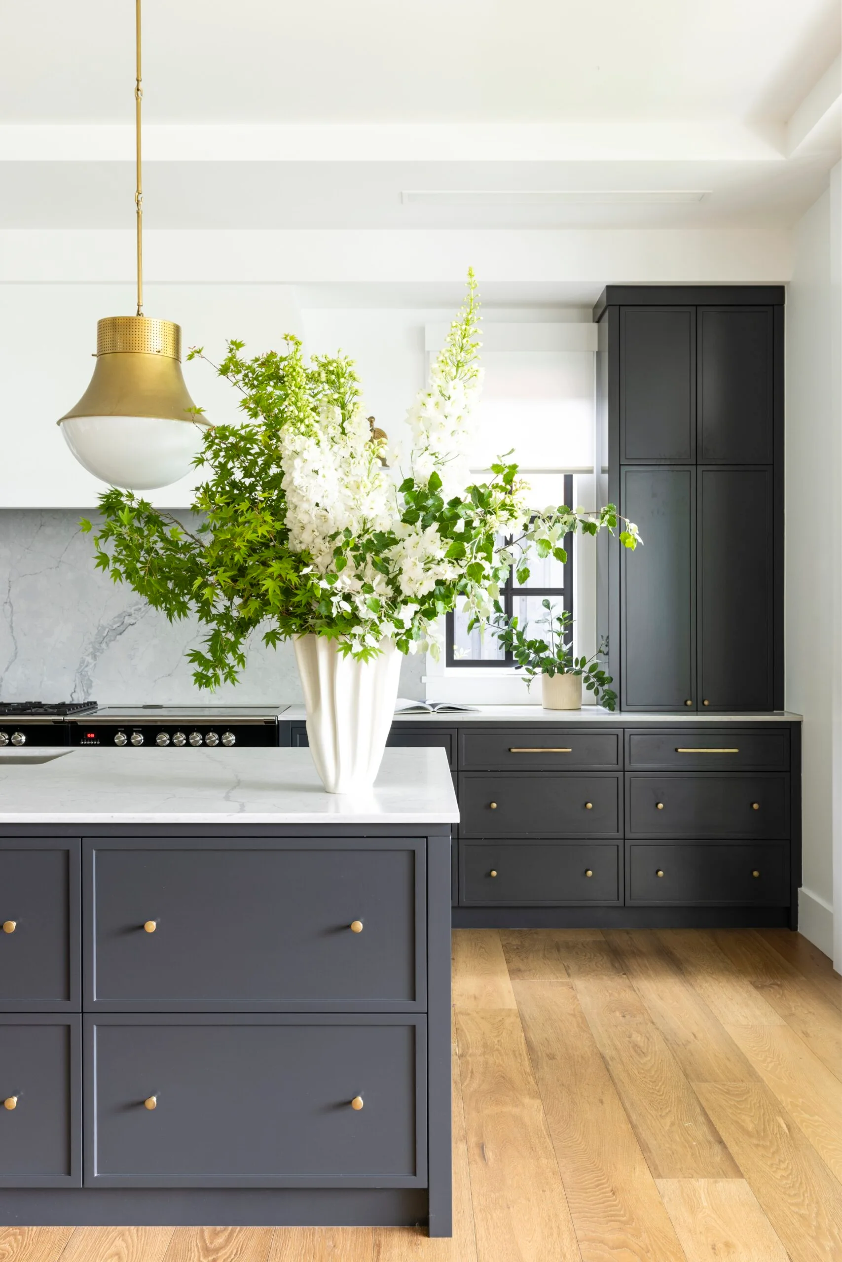Modern kitchen with dark grey cabinets with a shaker style profile, marble countertop, wood floor, pendant light, and a white floral arrangement on the island.