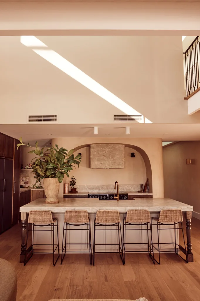 Spacious kitchen with a large marble island, wicker bar stools, potted plant, and sunlight streaming through a sunlight.