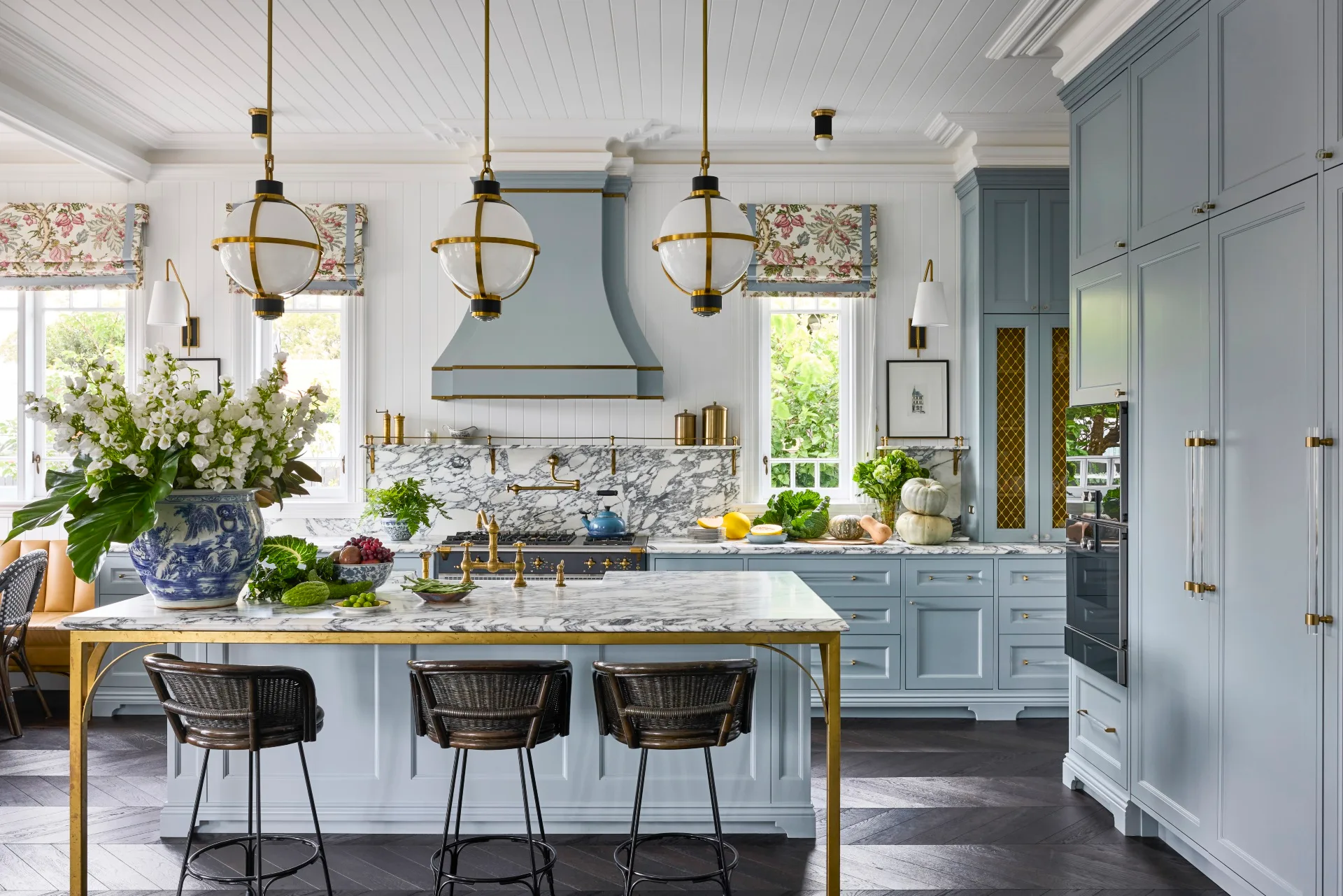 Elegant kitchen with blue cabinets, marble countertop and splashback, and brass fixtures. Large chinoiserie vase and pendant lights enhance charm.