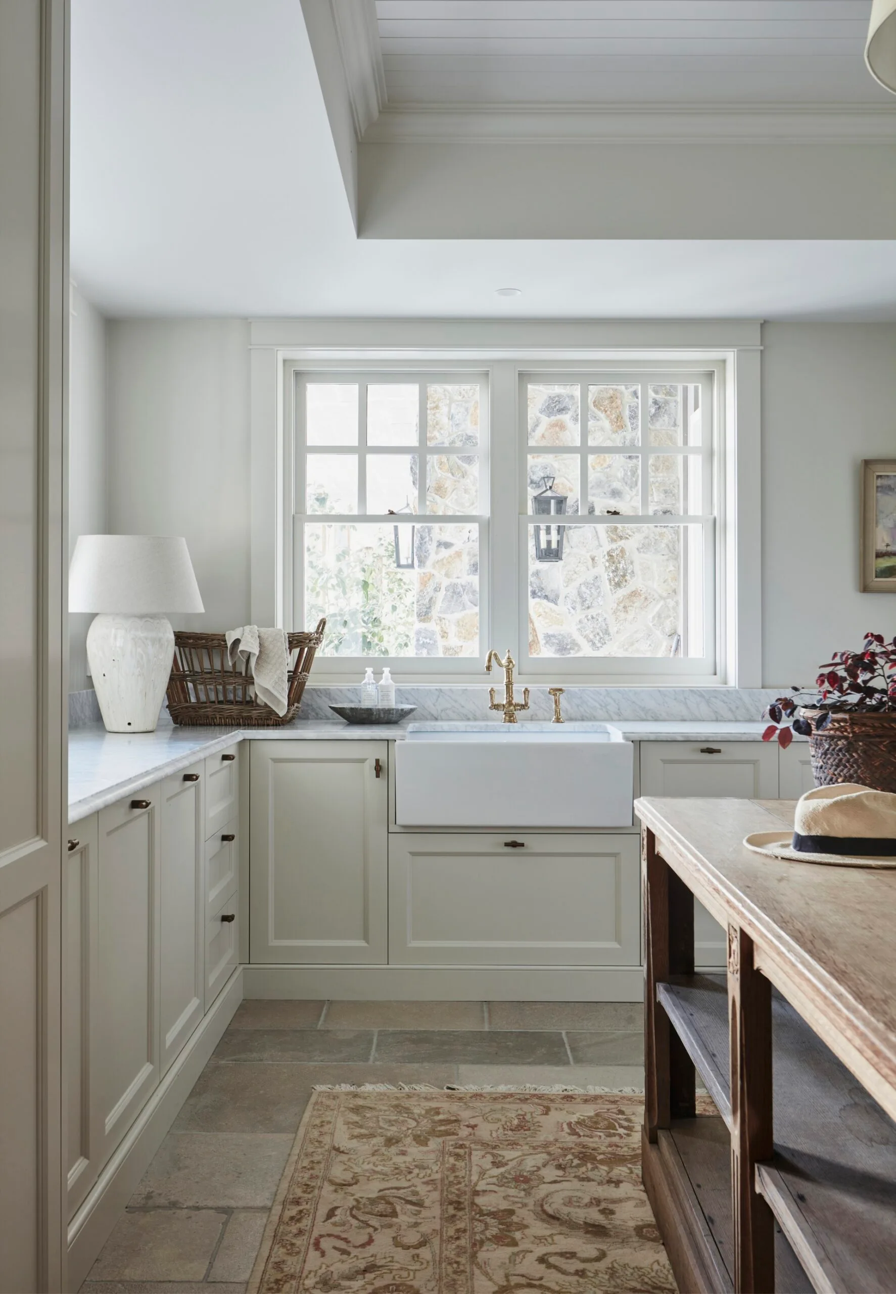 Bright kitchen with farmhouse sink, marble countertop, wicker basket, lamp, and wood island near a large window.