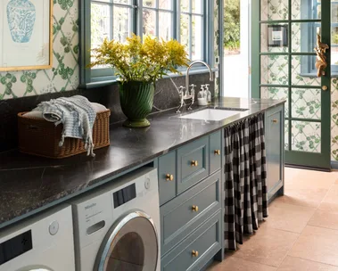 Laundry room with blue cabinets, green-patterned wallpaper, a vase of yellow flowers, and a washer and dryer and sink skirt