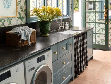 Laundry room with blue cabinets, green-patterned wallpaper, a vase of yellow flowers, and a washer and dryer and sink skirt