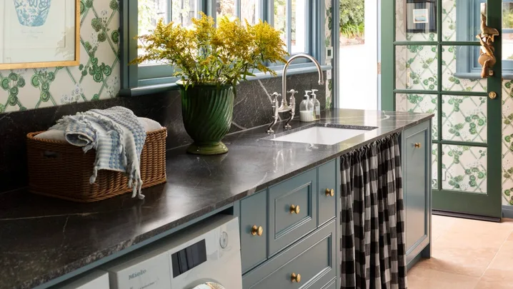 Laundry room with blue cabinets, green-patterned wallpaper, a vase of yellow flowers, and a washer and dryer and sink skirt