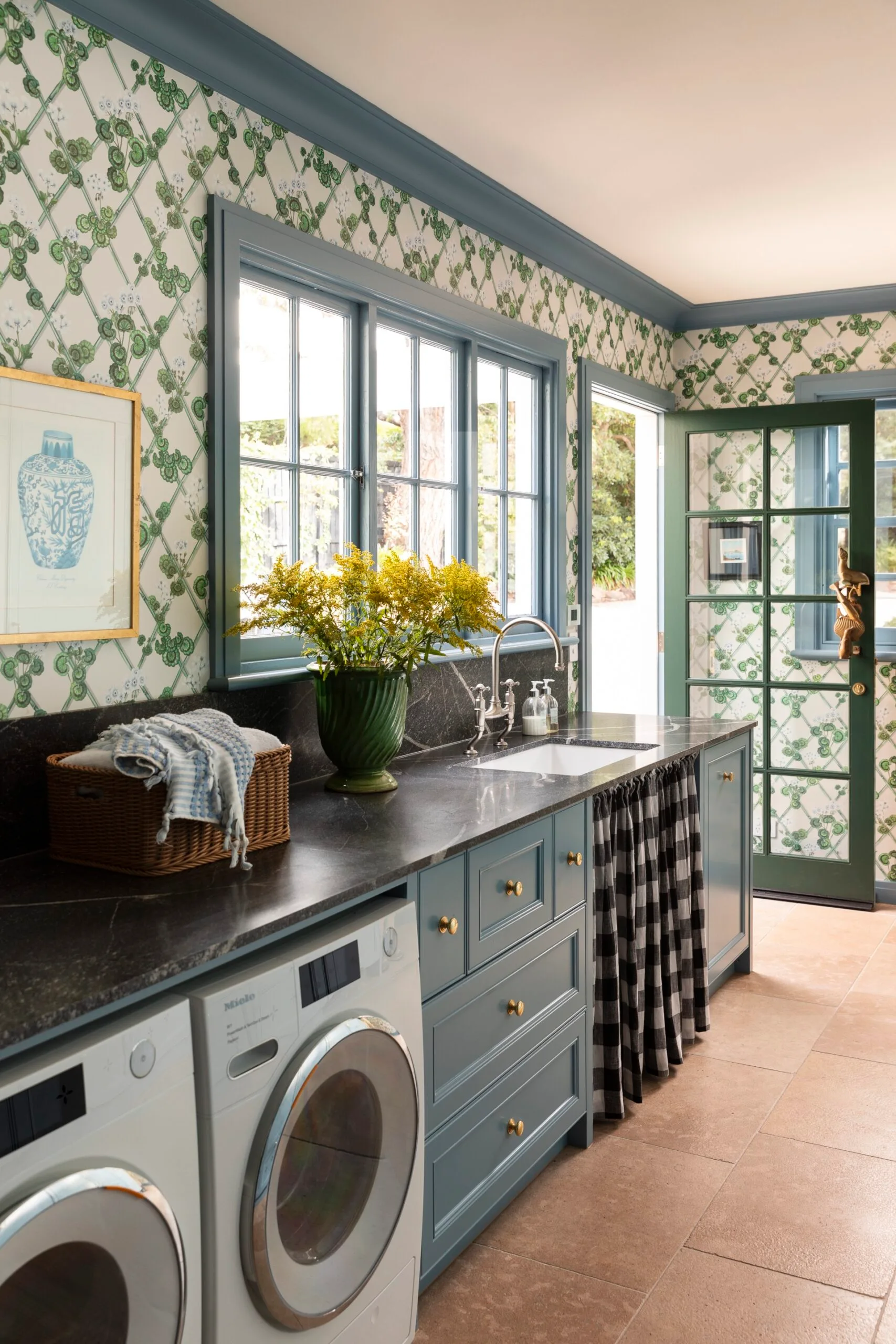 Laundry room with blue cabinets, green-patterned wallpaper, a vase of yellow flowers, and a washer and dryer.