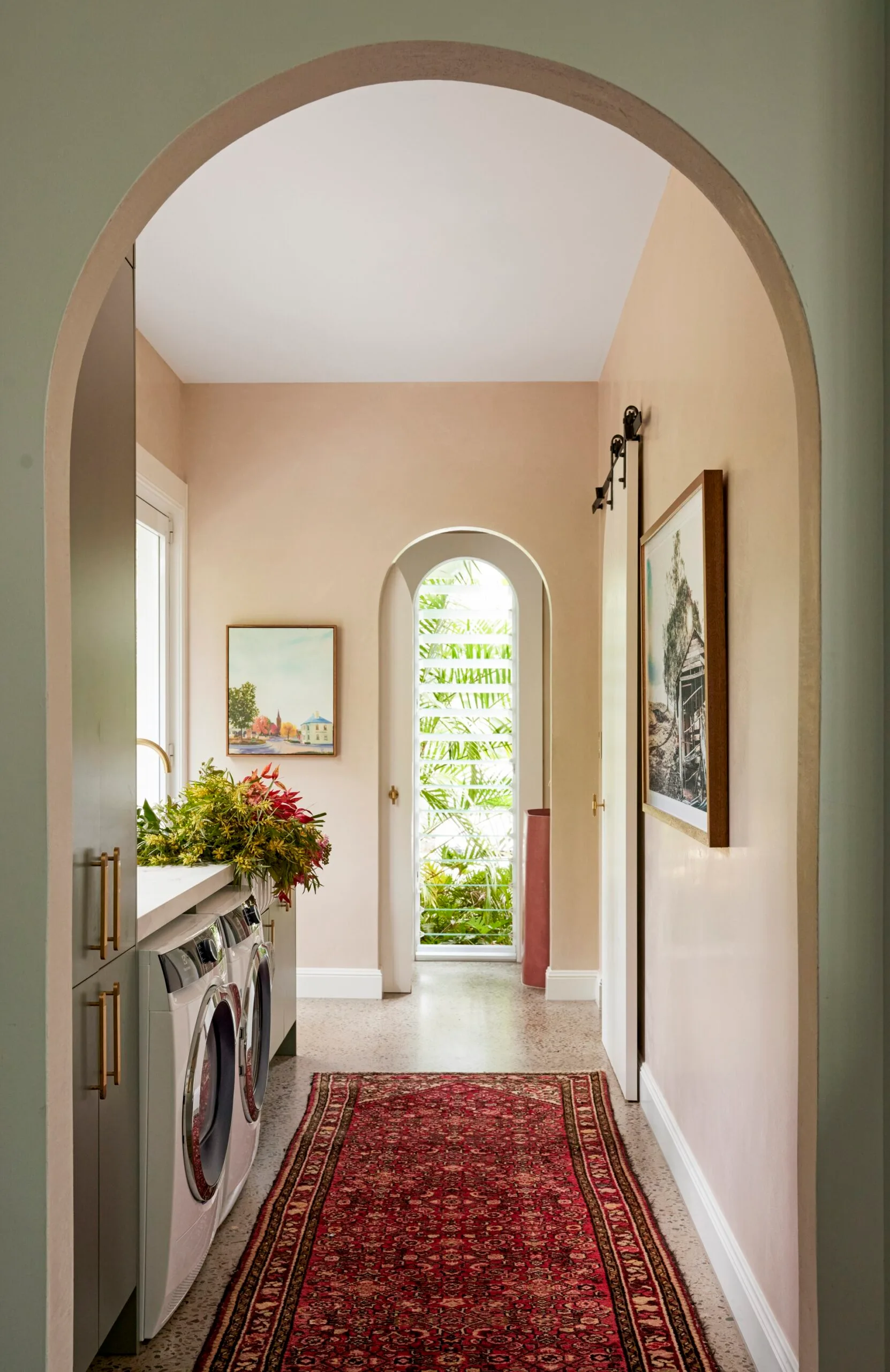 Stylish laundry room with arched doorways, elegant patterned rug, wall art, and plants, leading to a small glass door with garden view.