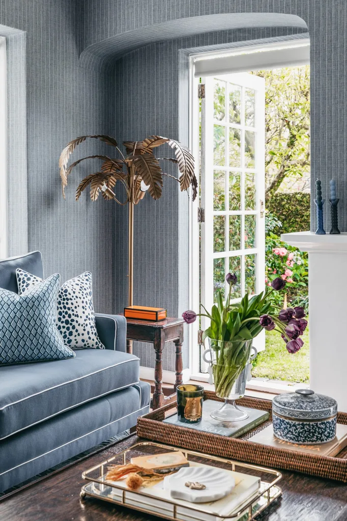 Cozy living room with blue sofa, patterned pillows, a floor lamp in the shape of a palm tree, open door to garden, and decorative items on table.