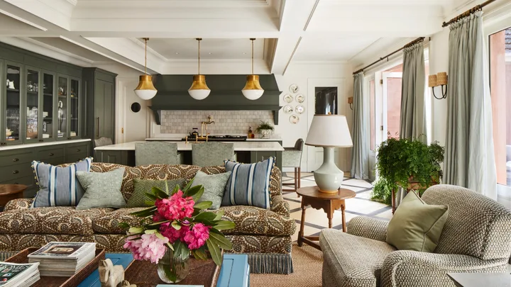Open-plan living room and kitchen with floral sofa, large windows, pendant lights, and green cabinets.