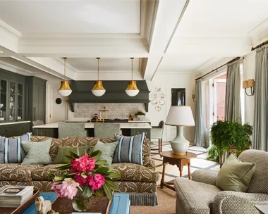 Open-plan living room and kitchen with floral sofa, large windows, pendant lights, and green cabinets.