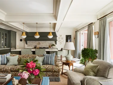 Open-plan living room and kitchen with floral sofa, large windows, pendant lights, and green cabinets.
