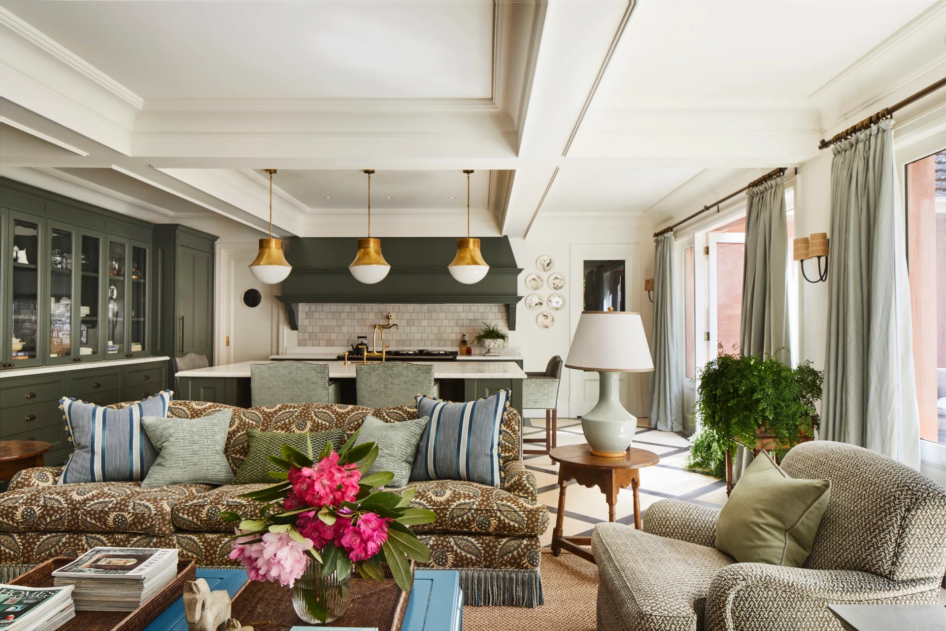 Open-plan living room and kitchen with floral sofa, large windows, pendant lights, and green cabinets.