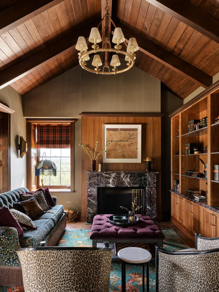 Cozy living room with vaulted wood ceiling, chandelier, floral sofa, leopard print armchairs, art, marble fireplace, and wooden bookshelf.