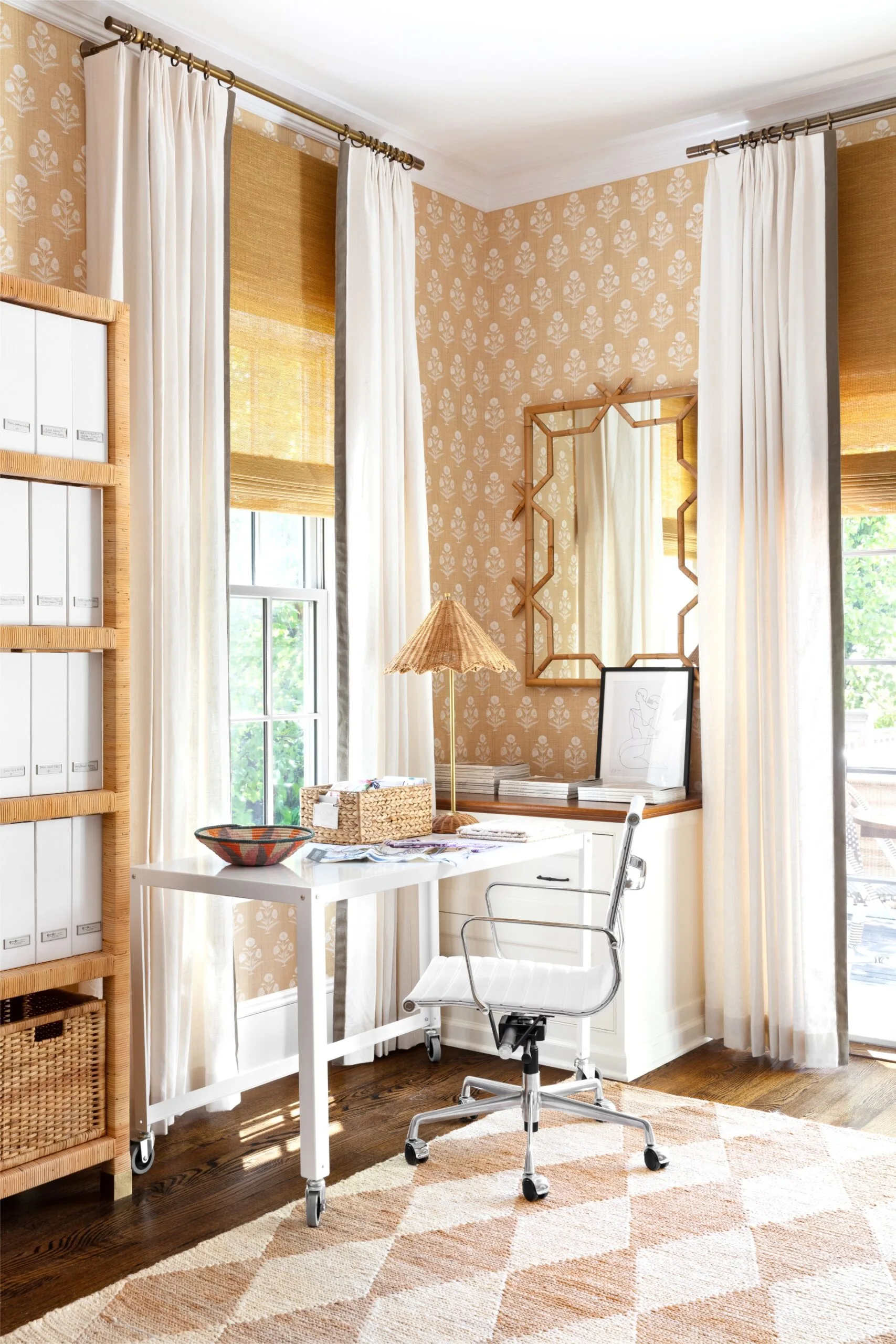 Cozy home office with a white desk, white and chrome desk chair, and patterned wallpaper. Natural light from large windows. The wallpaper and rug are cream and blush toned, with rattan boxes, table lamp and shelving unit.