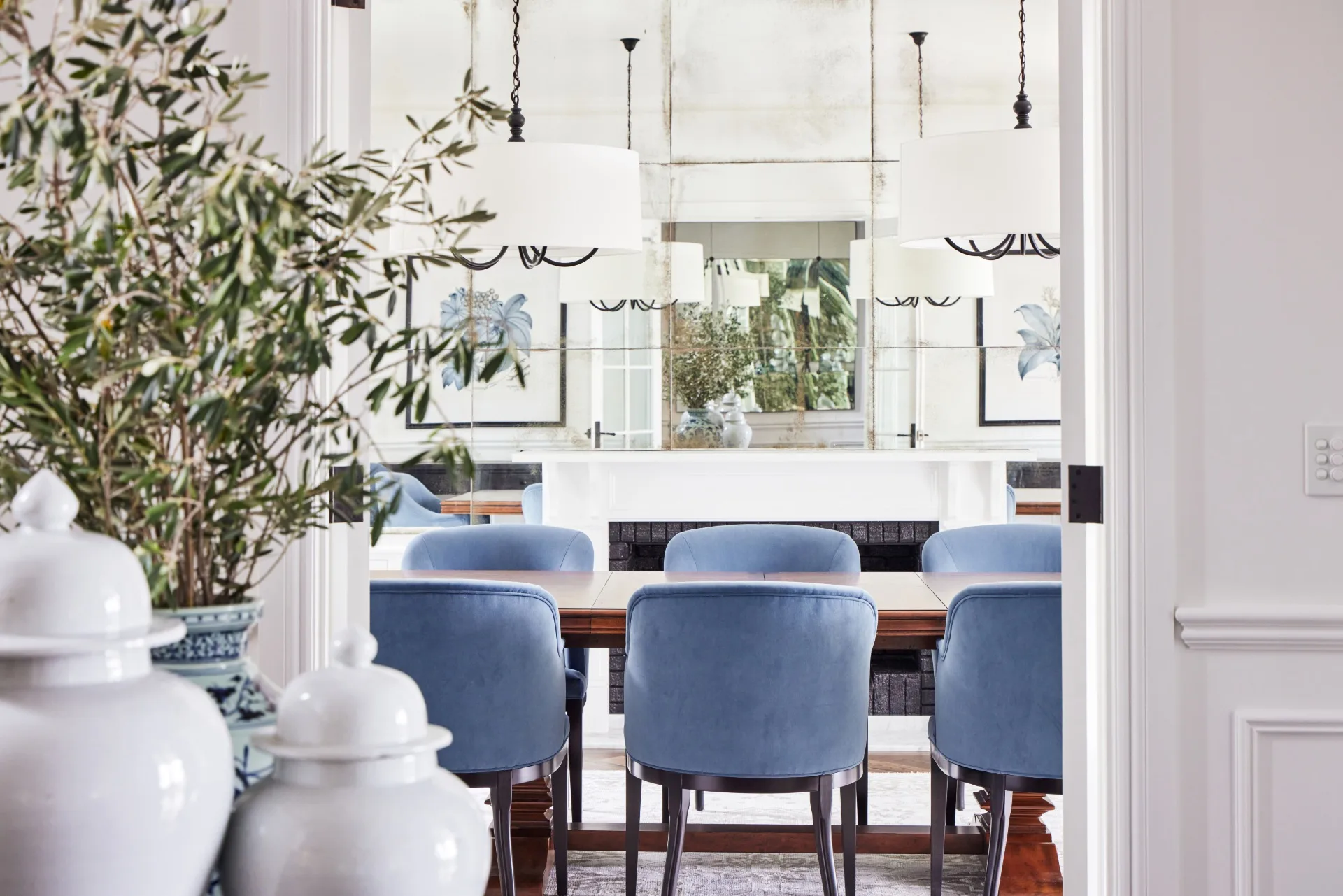 Dining room with blue chairs, white chandeliers, plants, and art on walls, seen through a doorway.